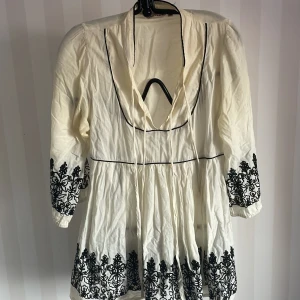Vit tunika med svart broderi från EastWest - Charmig vit tunika från EastWest i storlek 34. Den har vackra svarta broderier längs ärmarna och nederkanten. Tunika är långärmad och har en avslappnad passform med knytband vid halsen. Perfekt för vår och sommar! Legat i garderoben och i behov av tvätt , se bilder.
