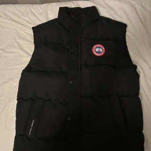 Svart dunväst från Canada Goose - Säljer en svart dunväst från Canada Goose i mycket bra skick. Västen har en hög krage och knappar framtill. Perfekt för kyliga höst- och vinterdagar. Den har en klassisk stil med det ikoniska märket på bröstet. Passar både för vardag och utomhusaktiviteter.
