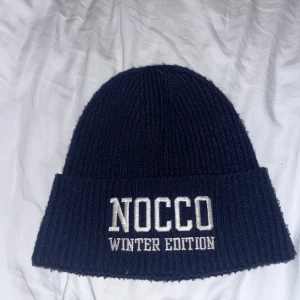 Mörkblå stickad mössa från NOCCO knappt använd - Säljer en snygg mörkblå stickad mössa från NOCCO Winter Edition. Den är perfekt för kalla dagar och har en broderad text framtill. Mössan är i mycket bra skick och passar perfekt för vintern. Håll dig varm och stilren! 