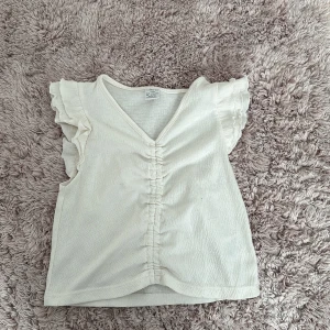 Vit ribbad topp med volangärmar - Säljer en söt vit ribbad topp med volangärmar och v-ringning. Perfekt för sommardagar eller en avslappnad vårlook. Toppen har en fin rynkdetalj framtill som ger den lite extra stil. Passar bra till både jeans och kjol!