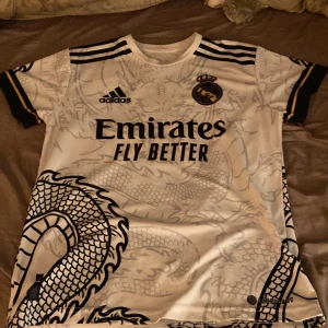 Vit Real Madrid tröja med drakmönster - Säljer en vit Real Madrid tröja från Adidas i storlek L. Den har ett coolt drakmönster i svart och guldiga detaljer. Kortärmad med 'Ronaldo 7' på ryggen och 'Hala Madrid!' tryckt i nacken. Perfekt för fans! ⚽