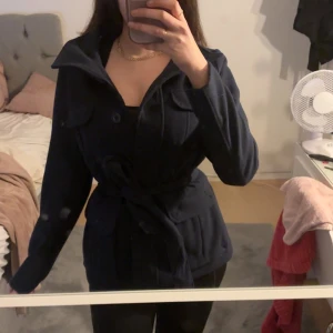 Mörkblå kappa från H&M - Säljer en snygg mörkblå kappa från H&M i storlek M 38 sitter som S på kroppen. Den har långa ärmar och ett bälte i midjan för en fin passform. Perfekt för höst och vår! Kappan har knappar framtill och en klassisk krage. . 