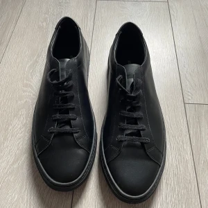 Original Achilles common projects - Säljer ett par stilrena svarta sneakers i skinn. De har en klassisk design med snörning och en diskret guldfärgad detalj på sidan. Sulan är i bra skick och de är perfekta för både vardag och fest. Passar till det mesta i garderoben!  44 pris kan diskuteras, köpt här på plick låda kommer med!