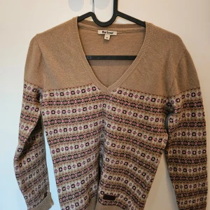 Beige mönstrad ull tröja från Barbour - Säljer en snygg beige ull tröja från Barbour med ett färgglatt mönster i rosa, svart och grönt. Tröjan har en v-ringad hals och långa ärmar, perfekt för kyligare dagar. Den är både stilren och bekväm.