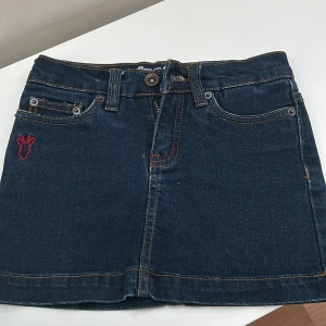 Jeanskjol från Baron Clothes - Snygg jeanskjol från Baron Clothes med klassisk femficksdesign och en liten röd broderad detalj framtill. Kjolen har en knapp och dragkedja i midjan och är perfekt för en avslappnad stil. Står för 6-7 år men passar till äldre ❤️