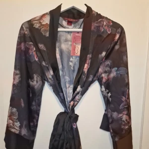 Kimono från Hunkemöller x Lucy Hale  - Sensuell kimono från Hunkemöller x Lucy Hale i siden. Den har mörka toner med rosa och vita blommor. Perfekt för en elegant och avslappnad look som inte säljes längre. Dyr i inköp. Garderobsrensning så kika gärna på övriga auktioner.