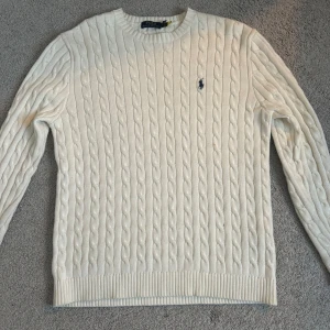 Vit stickad tröja från Ralph Lauren - Säljer en stilren vit stickad tröja från Ralph Lauren med kabelstickat mönster. Tröjan har långa ärmar och en rund halsringning. Perfekt för kyligare dagar!
