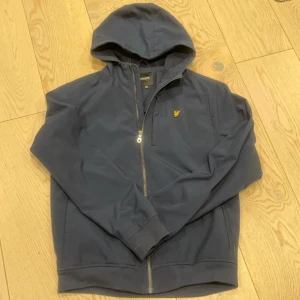 Mörkblå huvtröja från Lyle & Scott - Säljer en stilren mörkblå huvtröja från Lyle & Scott med dragkedja och en liten gul logga på bröstet. Perfekt för en avslappnad look med ribbade muddar och en bekväm huva.