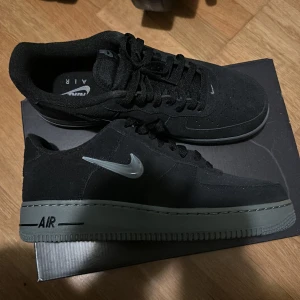 Svarta Nike Air Force sneakers - Snygga svarta Nike Air Force sneakers med en stilren design. Skorna har en svart ovandel med en grå sula och den ikoniska Nike-loggan i silver på sidan. Perfekta för en casual look.