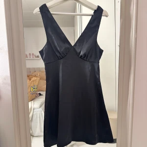 Svart klänning med djup urringning - Elegant svart klänning i satin med djup v-ringning. Perfekt för en stilren look. Klänningen har en smickrande passform och är ärmlös, vilket gör den idealisk för varmare väder.