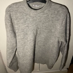 Grå stickad tröja från Zara - Säljer en stilren grå stickad tröja från Zara. Perfekt för kyligare dagar med sin mjuka och bekväma passform. Tröjan har långa ärmar och en klassisk rund halsringning.