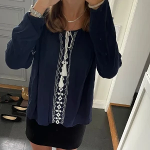 Mörkblå blus med broderi - Snygg mörkblå blus med vita broderade detaljer och knytning framtill. Blusen har långa ärmar och ett avslappnat snitt, perfekt för en bohemisk stil.