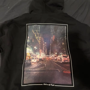 Svart hoodie med stadsmotiv - Snygg svart hoodie från Cav Empt med ett stort stadsmotiv på ryggen. Den har en liten vit logga på bröstet och en klassisk känguruficka framtill. Perfekt för en urban look! Helt ny!