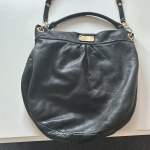 Svart axelväska från Marc by Marc Jacobs - Snygg svart axelväska i skinn från Marc by Marc Jacobs. Väskan har en elegant design med en metallplatta framtill och justerbara axelremmar. Perfekt för att bära dina dagliga nödvändigheter med stil.