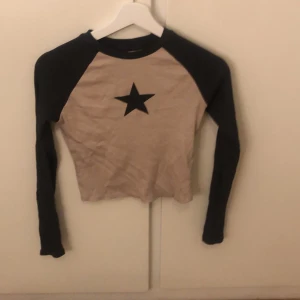 Beige och svart långärmad topp från H&M - Snygg beige topp med svarta ärmar och en stjärna på bröstet från H&M. Perfekt för en avslappnad stil med en cool touch. Långärmad design som passar bra till jeans eller kjol.