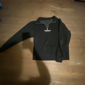 Svart tröja med dragkedja från Jack and Jones  - Snygg svart tröja från Jack and Jones  med halv dragkedja och vit text på bröstet. Perfekt för en avslappnad stil. Tröjan har långa ärmar och en bekväm passform.