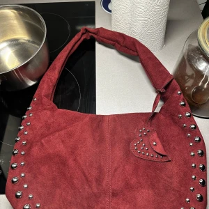 Röd axelväska med nitar - Snygg röd axelväska i mockaimitation med dekorativa nitar längs kanterna. Väskan har en praktisk storlek och ett hjärtformat hänge. Perfekt för att ge en edgy touch till din outfit. Inga defekter . Obs bilden visar inte riktigt färgen , den är mer vinröd i verkligen 