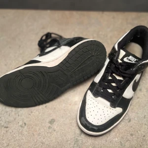 Nike sneakers i svart och vit - Säljer ett par svartvita Nike Dunk Low i storlek EU 38.5 (US 6Y). Skorna har en klassisk låg profil med perforerad tå och signatur Nike-logotyp på sidan. Perfekta för vardagsbruk eller streetwear-stil.  Skick: Begagnat skick.   Nypris: 1 100 SEK