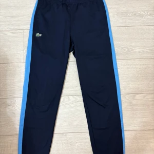 Lacoste Byxor - Snygga mörkblåa träningsbyxor från Lacoste med blå sidopaneler. Knappt använda och är i storlek S. Köpta på sandelins.