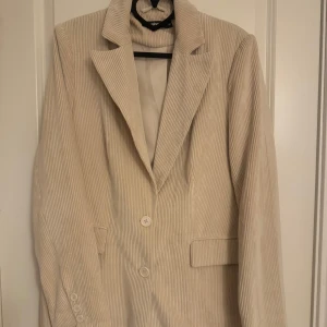 Beige manchesterkavaj - Gina Tricot - Snygg beige kavaj i manchester från Gina Tricot. Kavajen har en klassisk krage och knäpps med två knappar framtill. Perfekt för en stilren look.