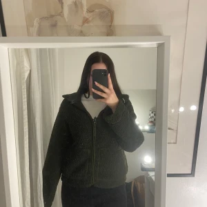 Grön teddyjacka med dragkedja - Mysig grön teddyjacka med dragkedja framtill. Perfekt för kyliga dagar med sin fluffiga och varma design. Jackan har en hög krage och långa ärmar för extra komfort. (Den är helt oanvänd men har inte lappen kvar).