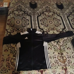 Svart träningsjacka från Adidas - Snygg svart träningsjacka från Adidas med vita detaljer och klassiska ränder på sidorna. Jackan har dragkedja och långärmade ärmar, perfekt för sportiga aktiviteter.