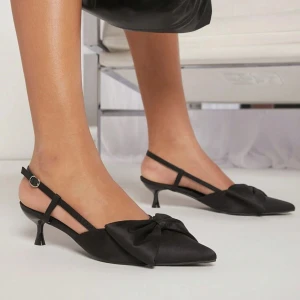 Svarta pumps med rosett - Eleganta svarta pumps med spetsig tå och en stor rosett framtill. Skorna har en smal rem runt hälen och en liten klack, vilket ger en stilren look. Perfekta för en chic och sofistikerad stil.