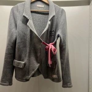 Grå kofta med rosa knytband - Mysig grå kofta från Odd Molly med rosa knytband framtill. Jackan har långa ärmar och två fickor. Perfekt för kyliga dagar.