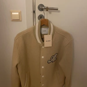 Beige bomberjacka från Axel Arigato - Snygg beige bomberjacka från Axel Arigato med broderad detalj på bröstet. Jackan har knappar framtill och en ribbad krage. Perfekt för en stilren look.