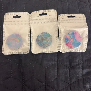 Färgglada runda pop sockets - Tre olika pop sockets med unika mönster i pastellfärger. En har ett mjukt regnbågsmönster, en annan har ett blått och grönt mönster som påminner om abalone, och den tredje har ett rosa och blått marmorerat mönster. Perfekt för att ge din telefon en personlig touch!