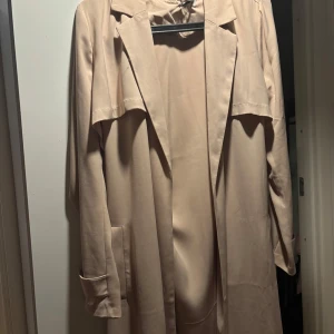Beige trenchcoat - Snygg beige trenchcoat med knytband i midjan och klassisk krage. Har en liten fläck