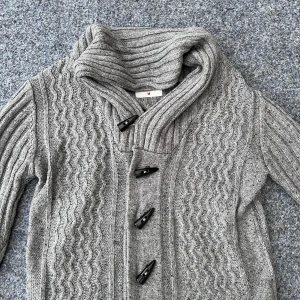 Grå stickad cardigan med knappar - Säljer en snygg grå stickad cardigan med ett vågigt mönster och stora svarta knappar. Nyskick med hög krage och långa ärmar, perfekt för vintern.