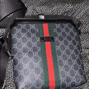 Svart axelväska med mönster och ränder - Snygg svart axelväska från Gucci med det klassiska GG-mönstret. Väskan har en grön och röd rand i mitten och en justerbar axelrem. Perfekt för att bära dina dagliga nödvändigheter med stil.