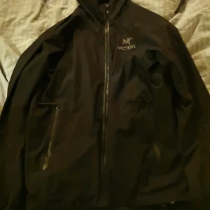 Svart jacka från Arc'teryx - Säljer en svart Arc'teryx jacka i Gore-Tex Infinium-material. Jackan har dragkedja och är perfekt för utomhusaktiviteter. Den är designad för att stå emot vind och väta, vilket gör den idealisk för varierande väderförhållanden. Har blivit använt som skid-jacka och delvit använt den för frilufs liv. Har ett lager fleece som isolation. Är öppen för frågor och priset är förhandlingsbart. 