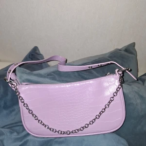 Lavendelfärgad axelväska med kedja - Fin lavendelfärgad axelväska i syntetmaterial med krokodil mönster. Väskan har en silverfärgad kedjedetalj och justerbar axelrem. Ett måttligt rymligt fack. 