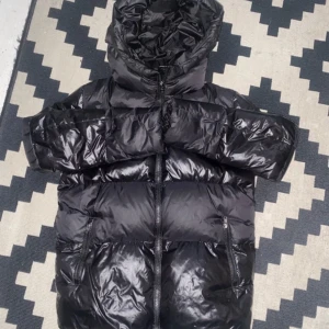 Intressekoll på min jacka - Säljer en svart dunjacka från Woolrich med huva och dragkedja. Jackan har en glansig yta och är perfekt för kalla dagar. Den har praktiska fickor med dragkedjor för säker förvaring. Det finns ett litet hål på ena armen. Högst 3500kr