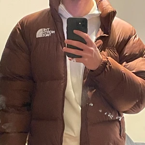 North Face 1996 Retro Nuptse Jacket - Säljer en brun dunjacka från The North Face med dragkedja och hög krage. Jackan har en pufferdesign och är perfekt för kyligare väder. nypris: 3900 kr. DM för funderingar, pris kan diskuteras.