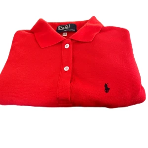 Röd pikétröja från Polo - Snygg röd pikétröja från Polo i 100% bomull. Tröjan har en klassisk krage och tre knappar framtill. Perfekt för en stilren look.