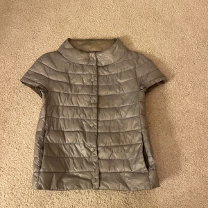 Grå kortärmad pufferjacka - Snygg grå pufferjacka med korta ärmar och knappar framtill. Perfekt för vår, sommarkvällar eller höst. Jackan har en gul fläck på baksidan, syns på bild tre och fyra. Vissa av knapparna på framsidan är dessutom lite sönder. Inget märke.