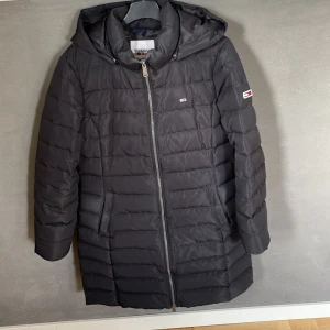 Svart pufferjacka från Tommy Jeans - Säljer en snygg svart pufferjacka från Tommy Jeans. Den har en praktisk huva och dragkedja framtill. Jackan är perfekt för kalla dagar och har en stilren design med Tommy Jeans-loggan på ärmen. Den är i storlek L och passar perfekt för vintern.