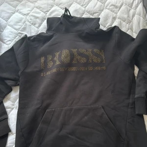 Svart hoodie från Boss - Säljer en svart hoodie från Boss med ett coolt  mönster på framsidan. Den har en hög krage och ribbade muddar vid ärmsluten. Perfekt för en avslappnad stil!