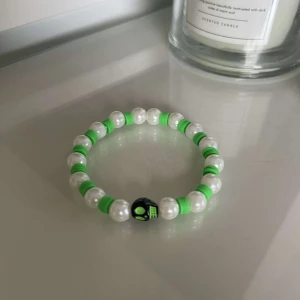 Pärlarmband med gröna detaljer - Säljer ett coolt armband med vita pärlor och gröna detaljer. Det finns också en svart pärla med ett grönt tryck som ger en unik touch. Perfekt för att piffa upp vilken outfit som helst!