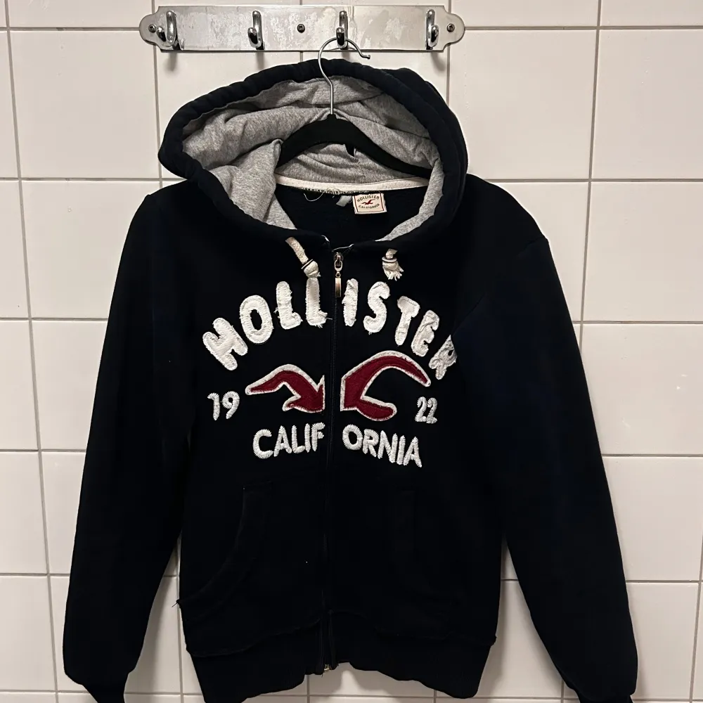 Säljer en mörkblå hoodie från Hollister med grått foder i luvan. Den har en dragkedja framtill och stora vita bokstäver med texten 'Hollister California' och årtalet 1922 i rött och vitt. Perfekt för en avslappnad stil! Har lite effekter på luvan men inget som påverkar tröjan. Osäker på storlek men skulle säga mellan XXS-S. Skriv för fler bilder eller frågor!💙🖤❤️🤍. Hoodies.