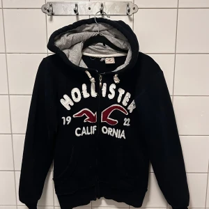 Svart hoodie från Hollister - Säljer en mörkblå hoodie från Hollister med grått foder i luvan. Den har en dragkedja framtill och stora vita bokstäver med texten 'Hollister California' och årtalet 1922 i rött och vitt. Perfekt för en avslappnad stil! Har lite effekter på luvan men inget som påverkar tröjan. Osäker på storlek men skulle säga mellan XXS-S. Skriv för fler bilder eller frågor!💙🖤❤️🤍