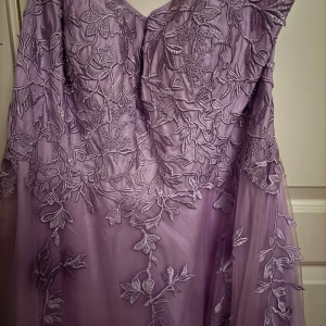 Ljuslila balklänning  - Helt oanvänd ljuslila näst intill syrenlila. Den har ett blommönster som är på överdelen av klänningen men sträcker sig lite till släpet. Den har snörning i ryggen och dragkedja. 