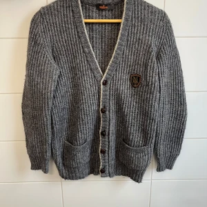 Grå stickad kofta från Morris - Säljer en snygg grå stickad kofta från Morris i storlek S. Den har en v-ringad design med knappar och två praktiska fickor fram. Koftan har också stiliga armbågslappar i beige och ett broderat emblem på bröstet. Perfekt för höst och vinter! Frågor och funderingar? Hör bara av dig