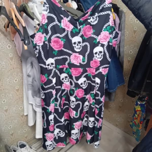 Svart klänning med dödskallar och rosor - Säljer en cool svart klänning med ett unikt mönster av vita dödskallar och rosa rosor. Klänningen har en v-ringad hals och är ärmlös, perfekt för en edgy look. Den är i normal passform och passar bra till både fest och vardag. Materialet känns mjukt och bekvämt.