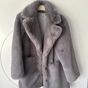 Grå pälsjacka från Chiqulle - Säljer en grå fusk pälsjacka från Chiquelle. Den är perfekt för kalla vinterdagar och har en klassisk design med långa ärmar och en stor krage. Jackan stängs med tryckknappar. Knapparna har lossnat & sömmarna i fickorna har släppt men de går att sy