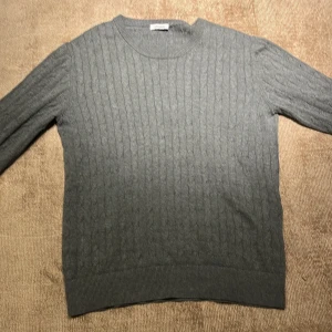 J.Lindeberg - Säljer en stilren grå stickad tröja från J.Lindeberg. Perfekt för kyliga dagar med sitt klassiska kabelstickade mönster. Tröjan har långa ärmar och en rund halsringning. Passar både till vardags och lite finare tillfällen. 🧶