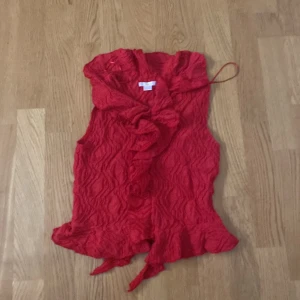 Röd volangtopp från Gina Tricot - Säljer en snygg röd topp från Gina Tricot med volangdetaljer. Toppen är ärmlös och har en vågig textur som ger en unik look. Perfekt för att liva upp garderoben med lite färg!